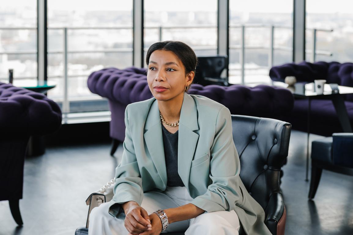 A confident woman in a mint blazer and gold chain necklace sits in a leather chair inside a stylish high-rise lounge with purple velvet sofas and a city skyline visible through large windows.
