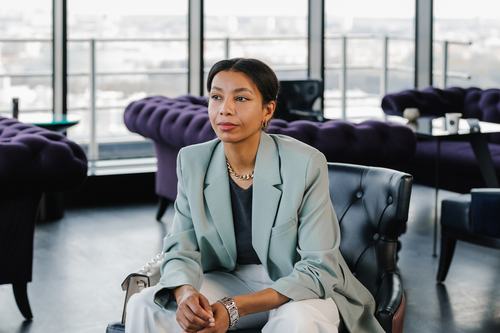 A confident woman in a mint blazer and gold chain necklace sits in a leather chair inside a stylish high-rise lounge with purple velvet sofas and a city skyline visible through large windows.