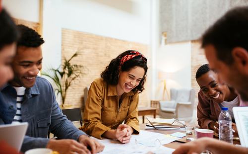 A diverse group of young people laugh and collaborate around a table covered with papers and notes in a bright, modern space. A smiling woman in a yellow jacket wearing a red headband is the focal point.