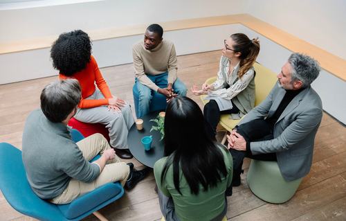 A diverse group of six people sit in a circle on colorful chairs around a small round table, engaged in conversation. The setting appears to be a modern office or collaborative workspace with wooden floors.