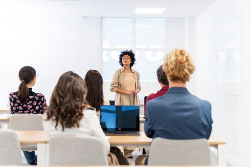 A woman with curly hair and glasses presents to a small group in a bright, modern classroom or office setting. Attendees sit at desks facing her, with a laptop open in the foreground and a projected display behind her.