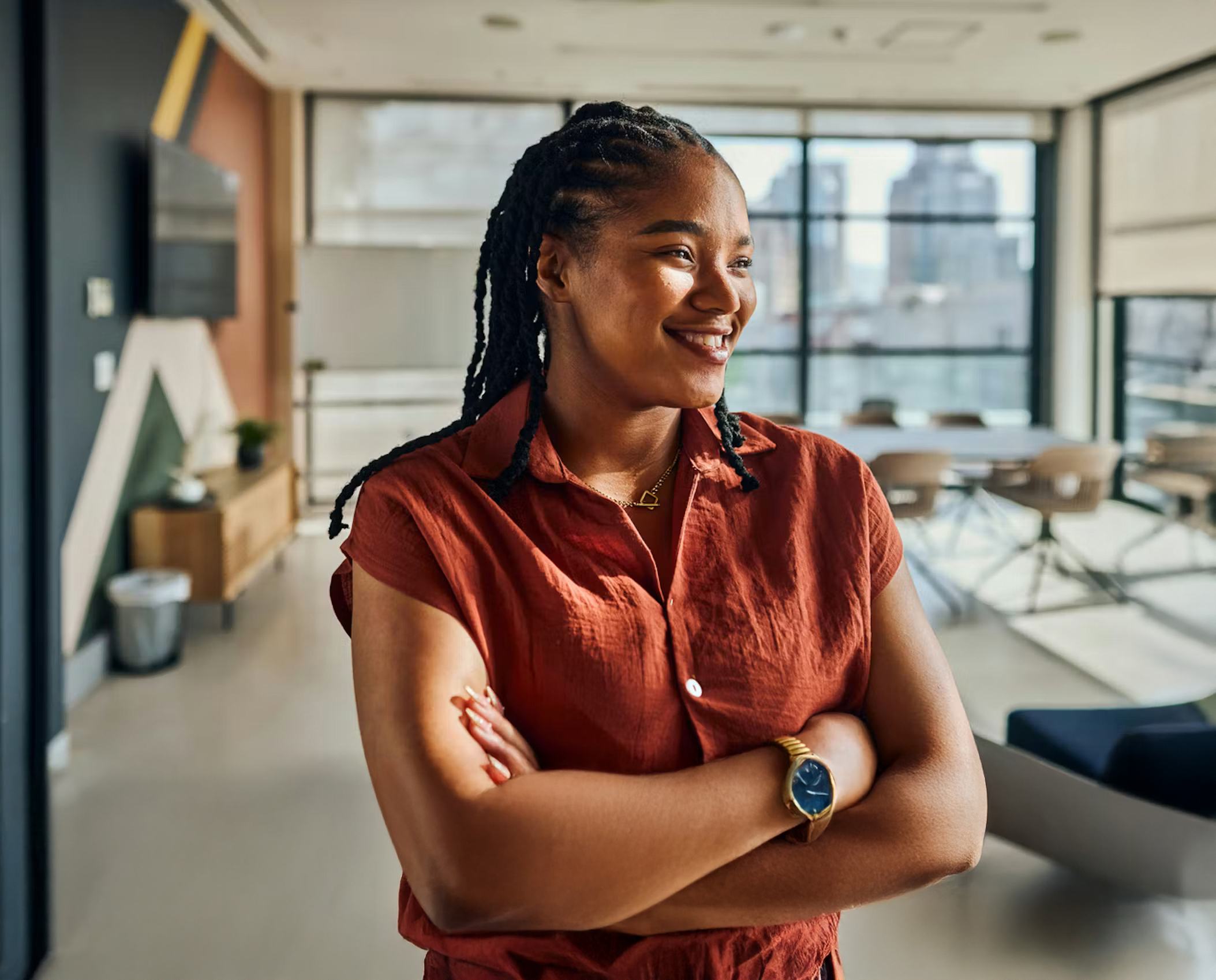 A confident young woman with braided hair smiles in a modern office, arms crossed, wearing a rust-red shirt, gold necklace, and watch. Bright windows and a meeting area are visible in the background.