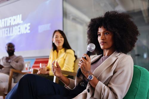 A woman with an afro speaks into a microphone at an Artificial Intelligence panel discussion, with two other panelists seated behind her and an AI-themed presentation slide visible in the background.