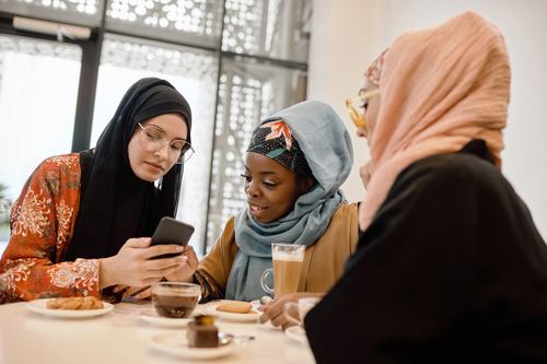 Three women wearing hijabs gather at a café table with coffee and snacks, looking at a smartphone together, sharing a warm, social moment.