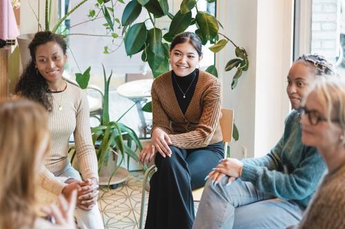A diverse group of women sitting together in a bright, plant-filled space, smiling and engaged in conversation. The relaxed setting suggests a casual meeting, support group, or friendly gathering.