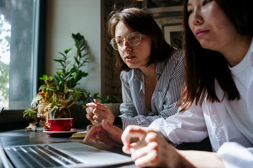 Two women collaborate at a café table with a laptop, a red coffee cup, and plants nearby. One woman wearing glasses and a striped shirt holds a pen, appearing to explain something to her colleague.