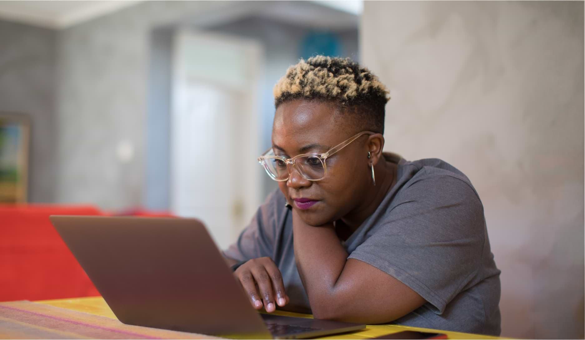 A woman with blonde-tipped natural hair and clear-framed glasses leans on a table, focused on her laptop screen. She wears a gray t-shirt and hoop earrings, working in a bright, modern indoor setting.