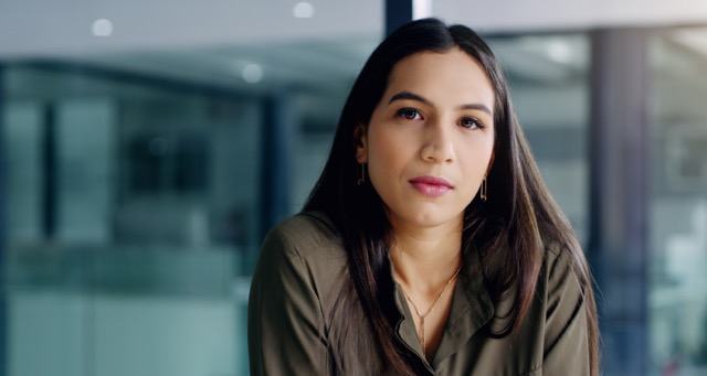A young woman with long dark hair wearing a olive green top and gold earrings, gazing confidently at the camera. She stands in a modern office environment with blurred glass walls in the background.