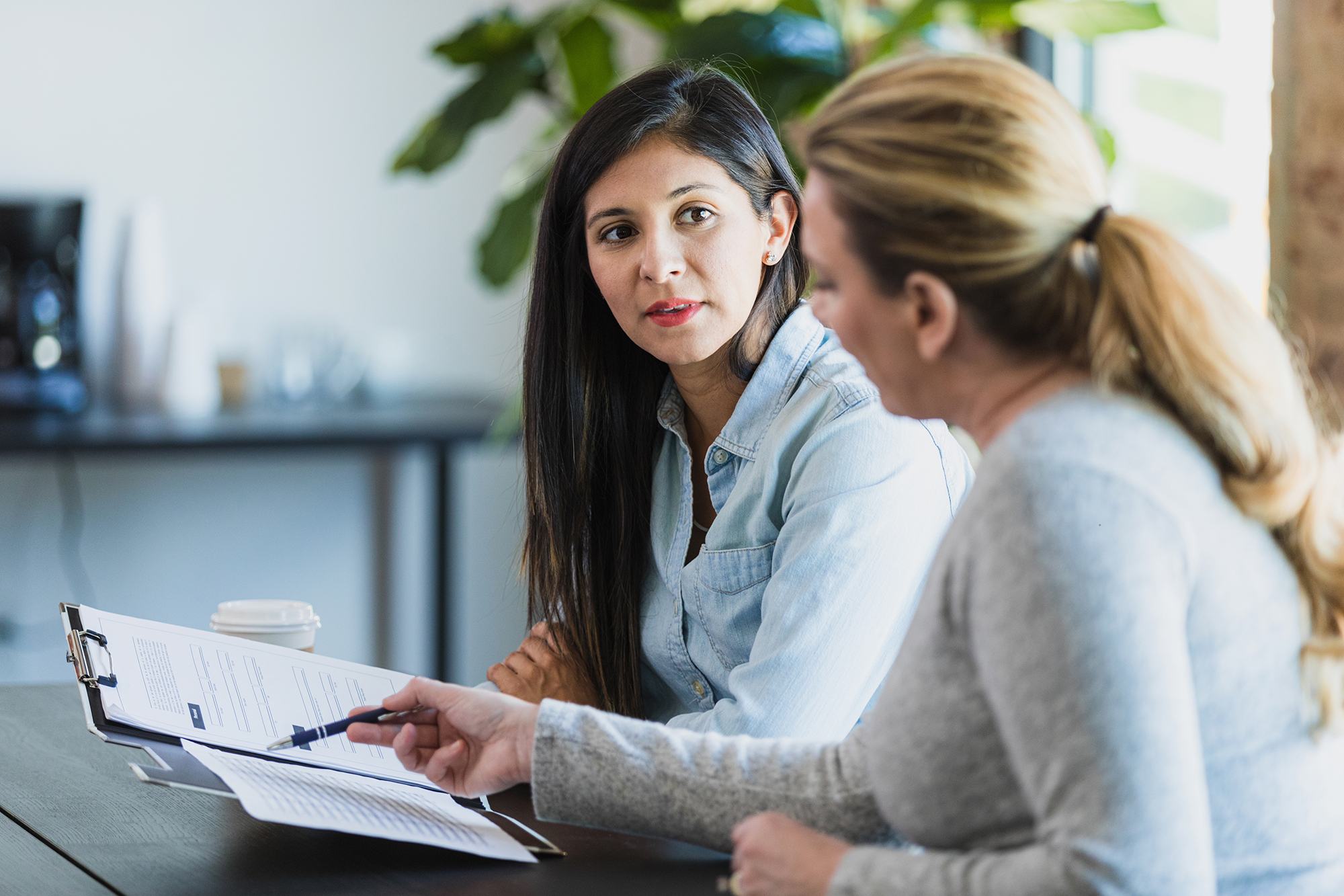 Two women sit at a table in a bright office, reviewing documents on a clipboard. One woman in a blue shirt listens attentively while the other points to the paperwork with a pen. A coffee cup sits nearby.