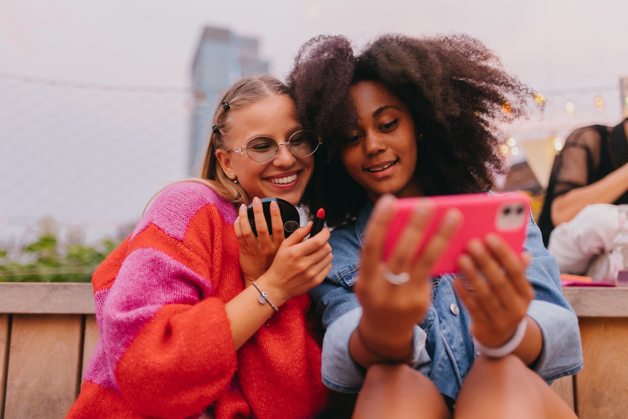 Two young women sit together outdoors, smiling as they look at a pink smartphone. One wears a colorful sweater and glasses; the other sports a denim jacket and curly hair. City lights and buildings are visible in the background.
