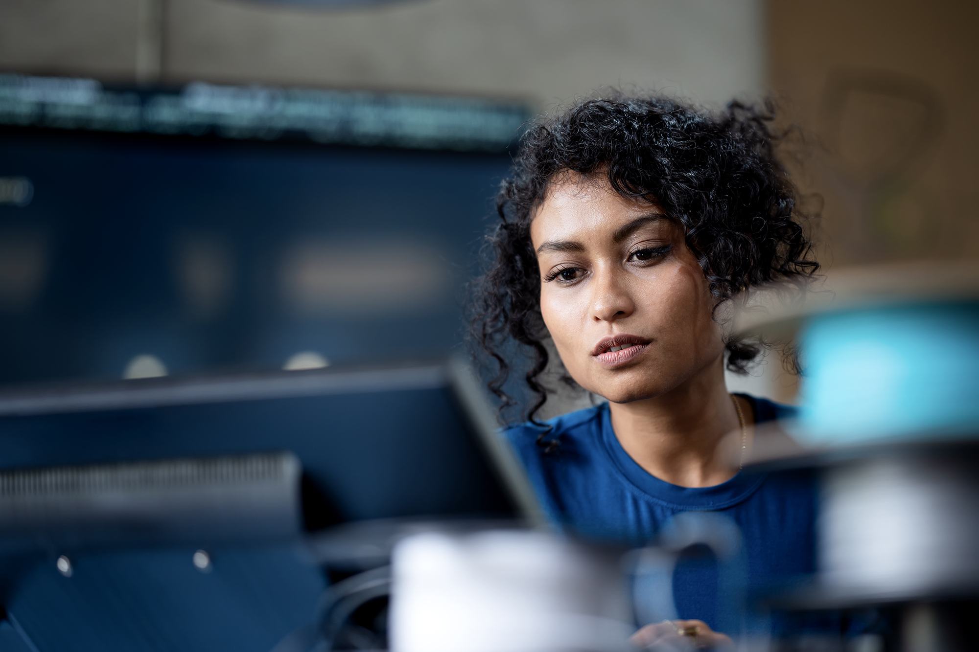 A young woman with curly hair, wearing a blue shirt, focuses intently on a computer monitor in an office or lab setting. The background is softly blurred, emphasizing her concentration.
