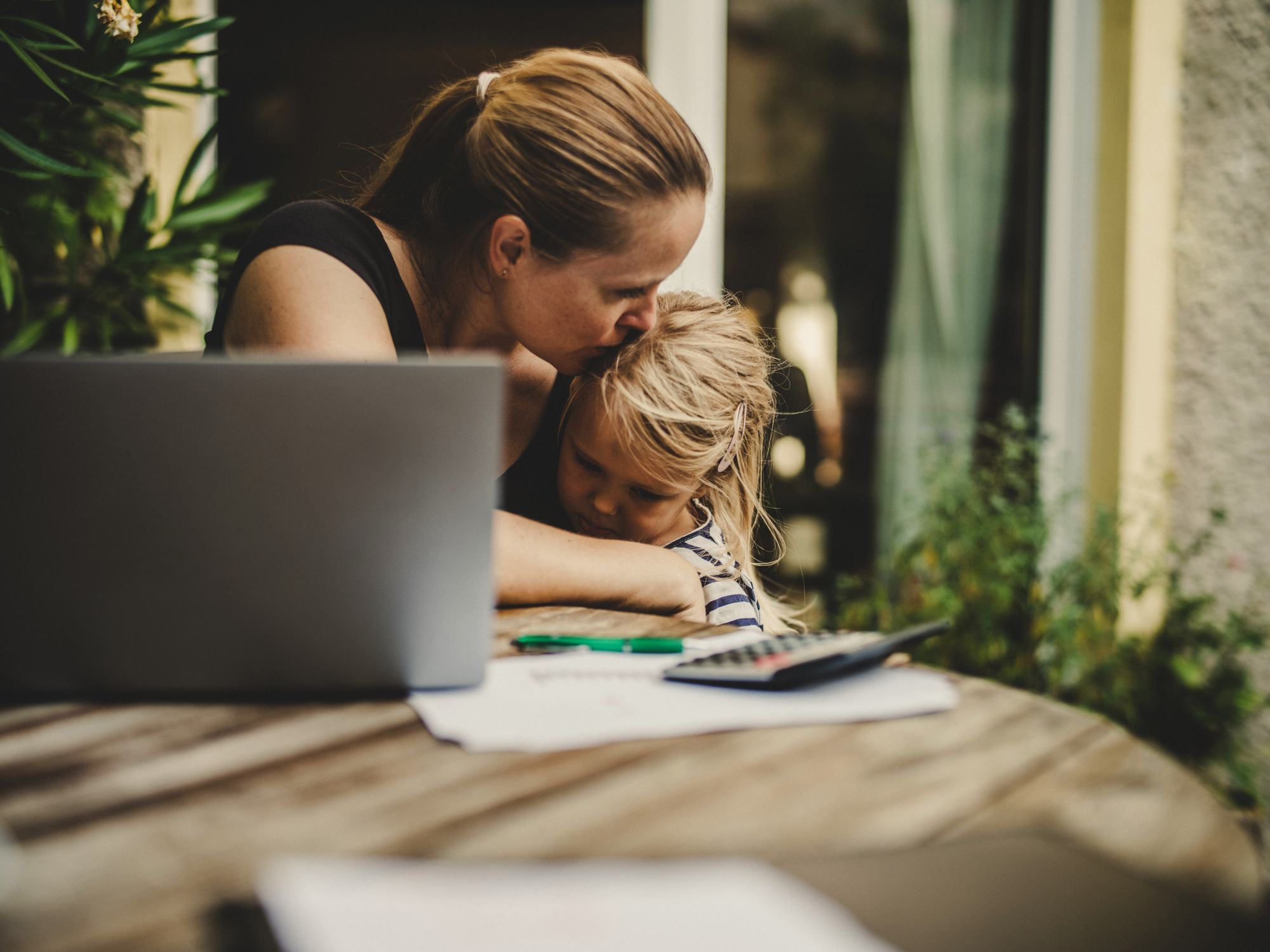 A mother kisses her young child's head while working at an outdoor table with a laptop, calculator, and papers, balancing parenting and remote work.