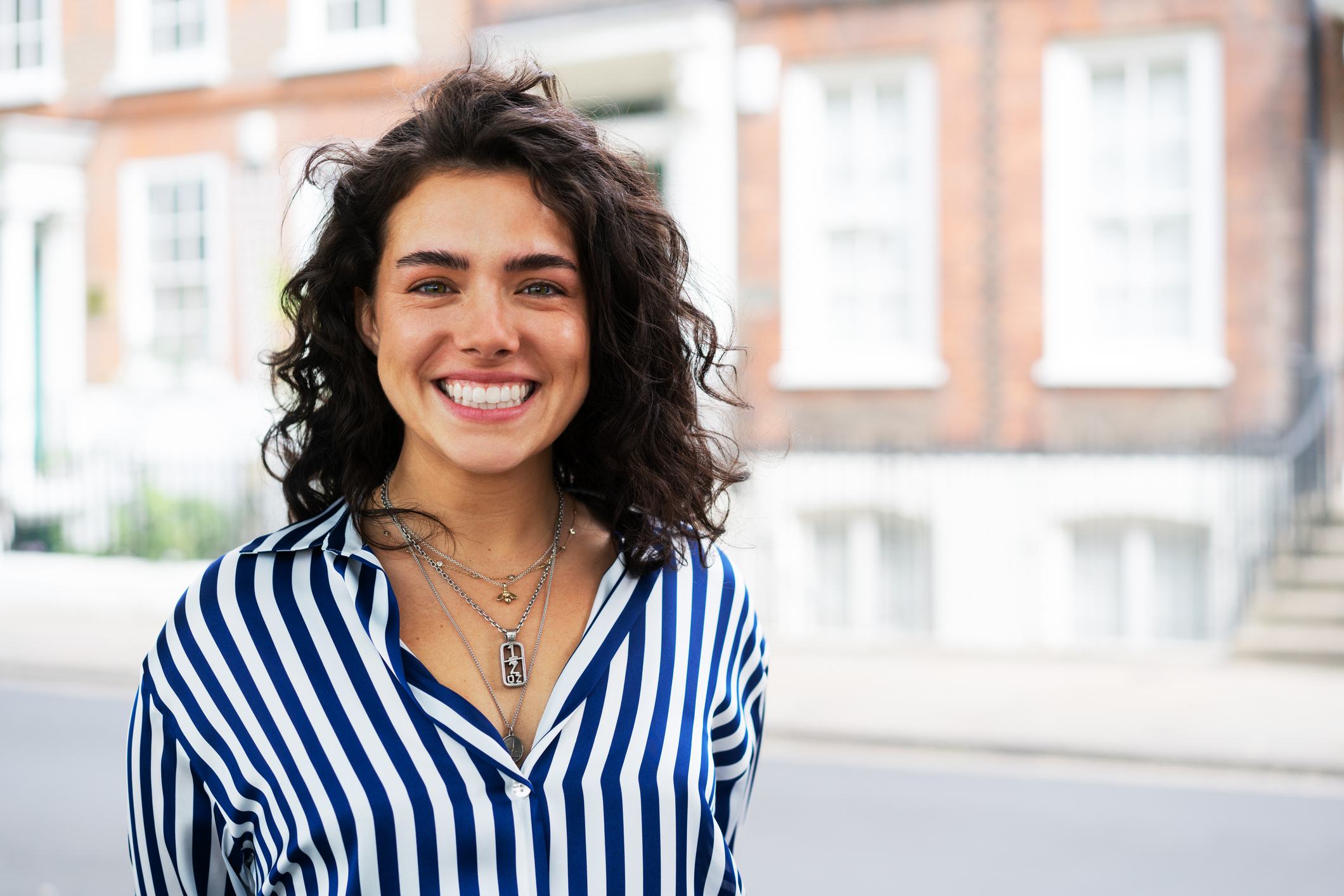 A young woman with curly dark hair smiles broadly outdoors, wearing a blue and white striped shirt and layered silver necklaces. A blurred brick building is visible in the background.