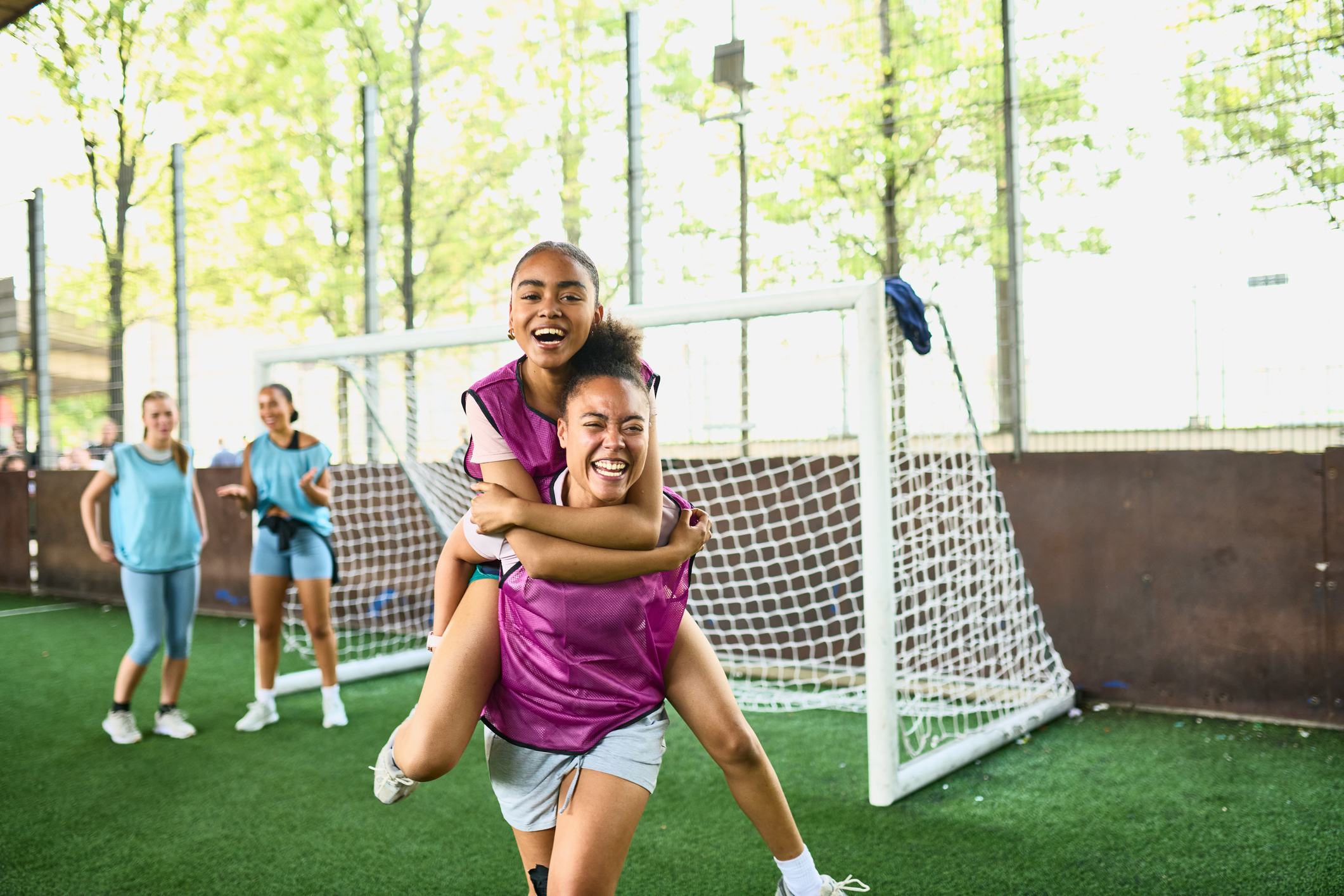 Two girls in purple vests celebrate joyfully on an outdoor soccer field, one giving the other a piggyback ride near the goal. Teammates cheer in the background on a sunny day.