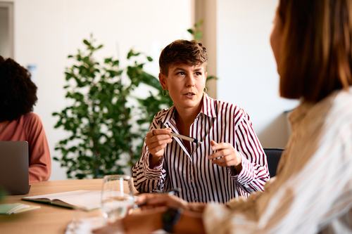 A person in a striped shirt holds glasses while speaking earnestly to a colleague across a table in a bright office setting, with a third person and a laptop visible in the background.