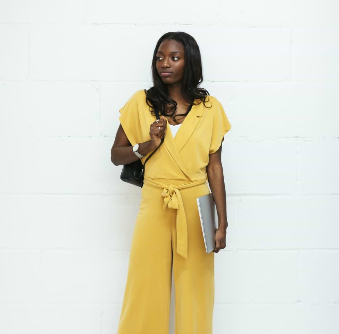 A stylish young woman wearing a mustard yellow wrap jumpsuit with wide-leg pants, holding a laptop, carrying a black bag, and wearing a watch, posed against a white brick wall.