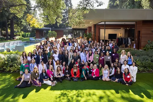 A large group of women pose for a photo on a lawn at an upscale outdoor venue with a pool and modern building. Many wear name badges, suggesting a professional conference or networking event on a sunny day.