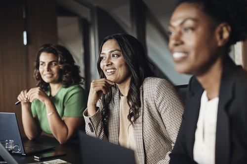 Three professional women smile and engage during a business meeting, seated at a conference table with laptops. They appear attentive and positive in a modern office setting.