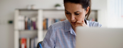 A focused woman in a striped shirt sits at a desk, pensively looking at a laptop while holding a pen, with a blurred bookshelf in the background.