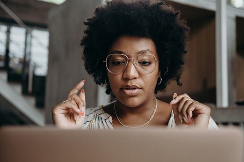 A young Black woman with an afro and aviator glasses looks intently at a laptop screen, hands raised as if explaining or thinking, wearing a gold necklace and colorful nail polish.