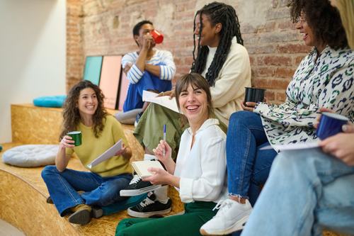 A diverse group of young people sit casually on wooden steps, laughing and chatting. They hold notebooks, pens, and colorful cups in a bright, brick-walled space, creating a relaxed, creative atmosphere.