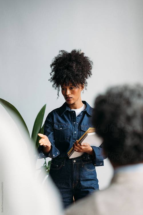A young woman with curly hair wearing a denim outfit holds books while speaking and gesturing expressively, likely presenting or teaching to an audience in a bright, minimalist setting with a plant nearby.
