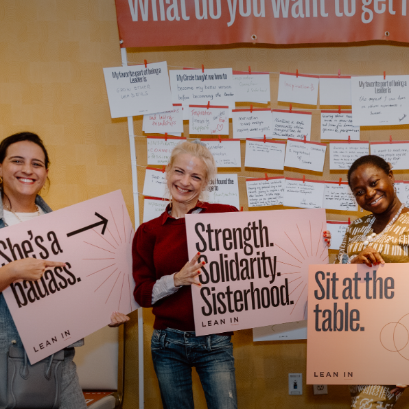 Three smiling women hold Lean In signs reading "She's a badass," "Strength. Solidarity. Sisterhood," and "Sit at the table." They stand before a board covered in sticky notes at what appears to be a women's empowerment event.
