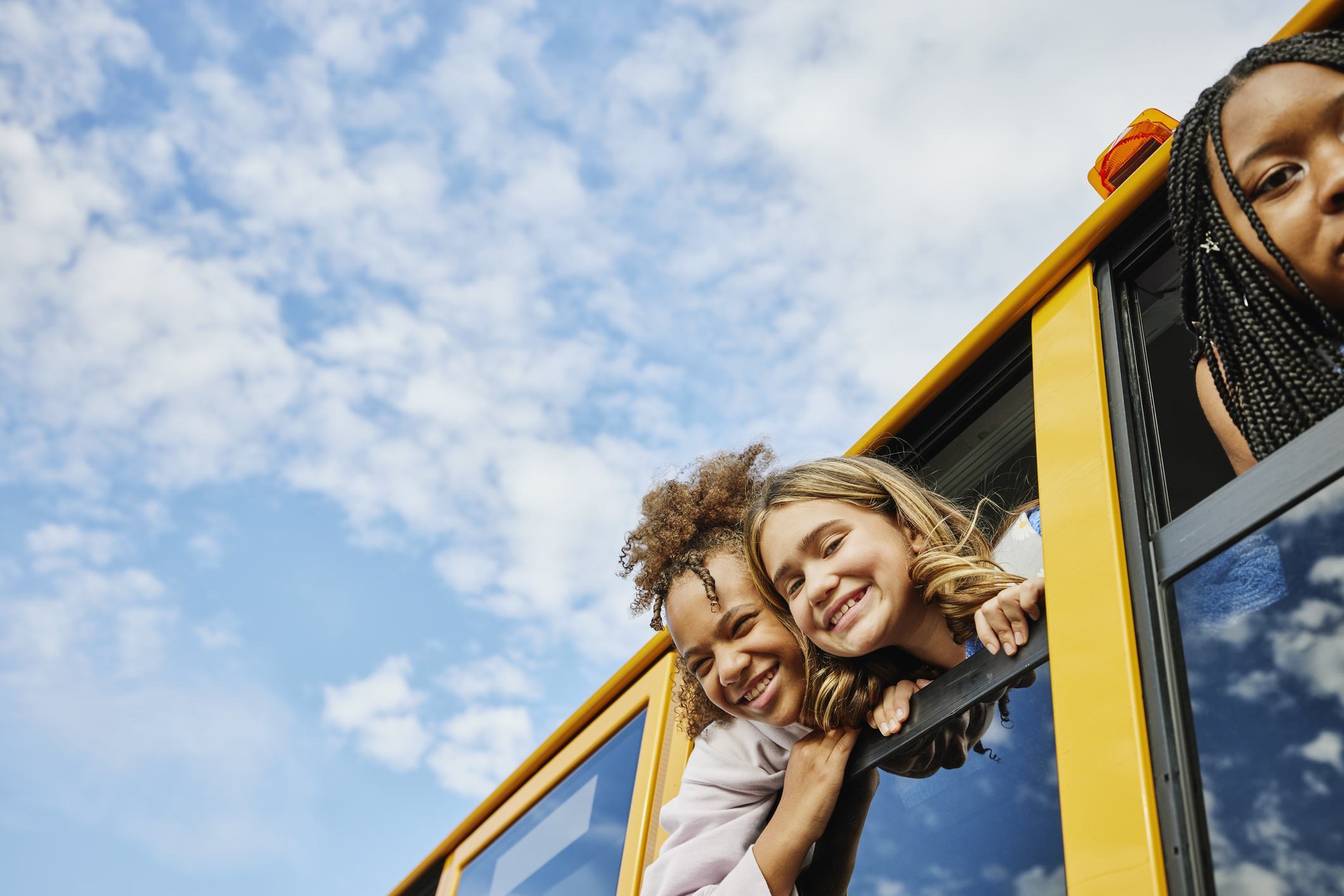 Three young girls smile as they lean out of a yellow school bus window against a bright blue sky, building confidence through friendship.
